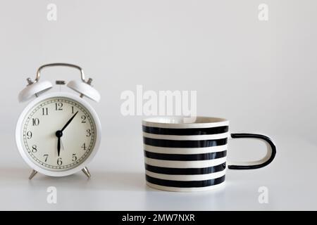 Tasse in schwarz-weißen Streifen auf einem weißen Tisch. Horizontaler Rahmen. Platz für Text. Ein weißer Wecker, ein sechs-Pointer Stockfoto