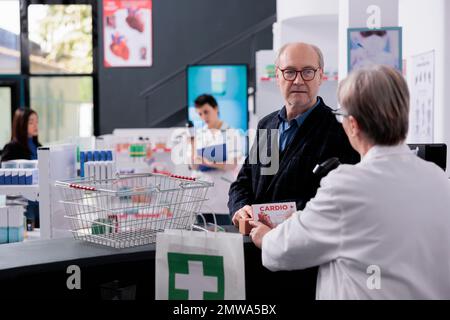 Leitender Kunde, der Herzvitamine an der Kasse einer Drogerie kauft und darauf wartet, dass ein Pharmaspezialist den Medikamenten-Barcode scannt. Älterer Mann mit Brille, der neben dem Schalter der Apotheke steht Stockfoto