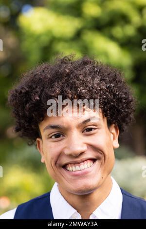 Vertikales Porträt eines lächelnden, birassischen Bräutigams mit lockigem Haar im Garten bei einer Hochzeit im Freien Stockfoto