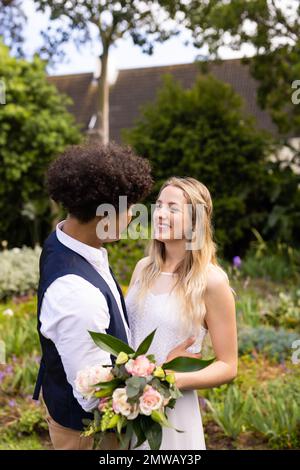 Eine Vertikale von glücklichen, vielseitigen Braut- und Bräutigams, die einen Blumenstrauß halten und bei einer Hochzeit im Freien lächeln Stockfoto