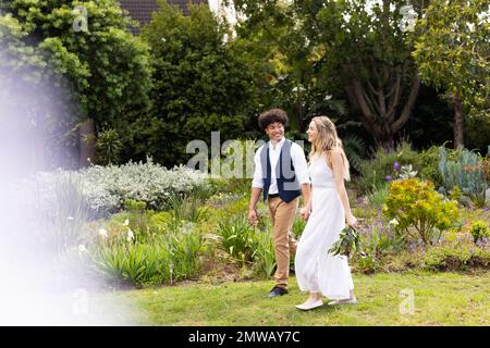 Glückliche, vielseitige Braut und Bräutigam, die Händchen halten, bei einer Hochzeit im Garten spazieren gehen, mit Kopierbereich Stockfoto