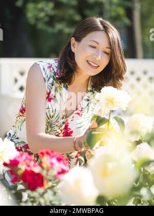 Eine weiße Frau bewundert blühende Rosen im öffentlichen Park. Sommerliche Stimmung. Tropische Pflanzen und Blumen blühen im Garten. Stockfoto