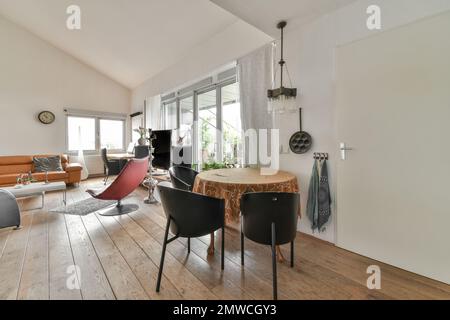 Ein Wohnzimmer mit Holzfußboden und weißen Wänden, einschließlich eines runden Esstisches, umgeben von zwei schwarzen Lederstühlen Stockfoto