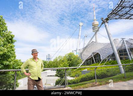 Freundlicher lächelnder Mann am Olympiaturm mit olympischem Zeltdach, BMW-Turm auf der linken Seite, Olympiapark, Olympiagelände, München, Oberbayern Stockfoto