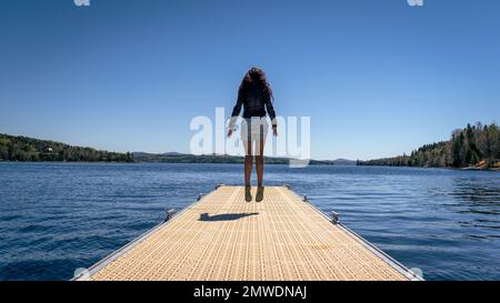 Nahaufnahme einer Frau, die auf dem Metalldock über dem See vor blauem Himmel springt Stockfoto