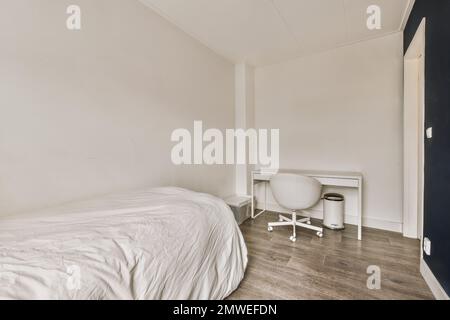 Ein Schlafzimmer mit Holzfußboden und weißen Laken auf dem Bett vor dem Zimmer ist ein leerer Schreibtisch Stockfoto