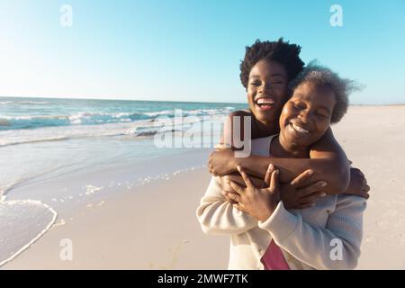 Fröhliche afroamerikanische junge Frau, die lacht und Mutter umarmt, während sie gegen Meer und Himmel steht Stockfoto