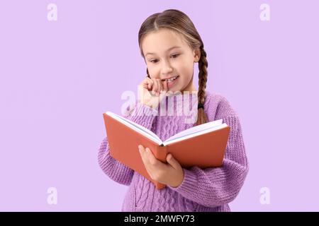 Ein kleines Mädchen, das Nägel beißt, während es ein Buch auf lila Hintergrund liest Stockfoto