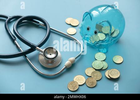 Konzept des Sparens für Krankenversicherungskosten oder Spesen- und Finanzkontrolle. Transparentes Sparschwein, Münzen und Stethoskop auf blauem Hintergrund. Stockfoto