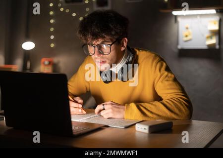 Attraktiver arabischer Student, der auf einem Laptop lernt, Notizen in einem Notizbuch schreibt und nachts im Schlafsaal sitzt. Speicherplatz kopieren Stockfoto