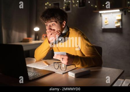 Schockierter junger Mann mit Laptop-Computer mit leerer Kreditkarte, der nachts zu Hause am Tisch saß. Speicherplatz kopieren Stockfoto