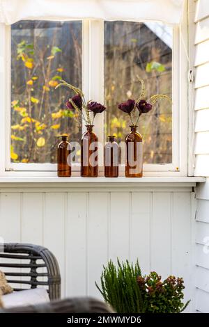Ein Set brauner dekorativer Flaschen, die als Vasen für wunderschöne künstliche Blumen am Fenster dienen Stockfoto