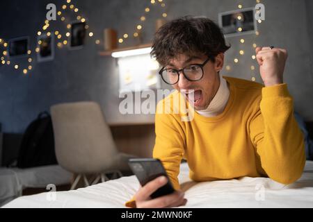 Überrascht, glücklicher junger Mann, der ein Handy hält und auf das Handy schaut, hat gute Nachrichten in sms gelesen, lag im Bett und war begeistert von der App WIN Scream wi Stockfoto