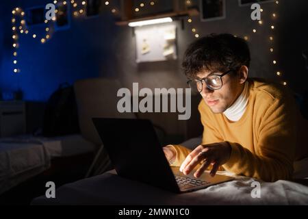 Glückliche junge Schülerin mit Brille, die nachts online lernt, Nachrichten liest, sich ein Schulungs-Webinar ansieht, das zu Hause im dunklen Raum auf dem Bett liegt Stockfoto