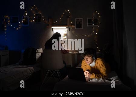 Männlicher Student, der Kopfhörer trägt, liegt auf dem Bett in einem gemeinsamen Haus und arbeitet an einem Laptop und einem Mädchen, während er im Hintergrund lernt. Speicherplatz kopieren Stockfoto