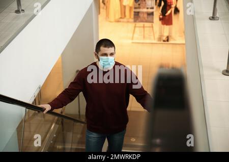 Mann mit Einwegmaske auf Rolltreppe im Einkaufszentrum. Gefährlicher Virus Stockfoto
