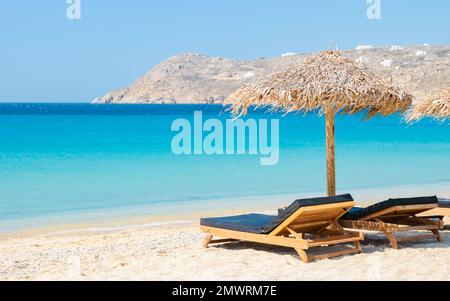 Mykonos Strand im Sommer mit Sonnenschirm und luxuriösen Liegestühlen, blauer Ozean mit dem Berg am Elia Beach Mikonos Griechenland. Stockfoto