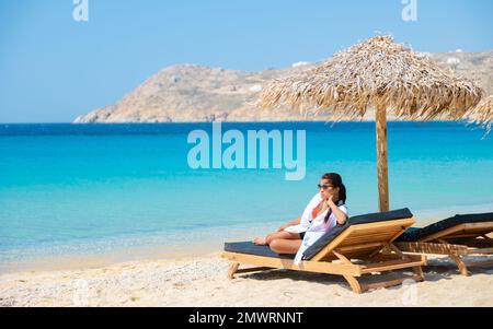 Frauen am Mykonos Strand im Sommer mit Sonnenschirm und luxuriösen Liegestühlen, blauer Ozean mit dem Berg am Elia Beach Mikonos Griechenland. Stockfoto