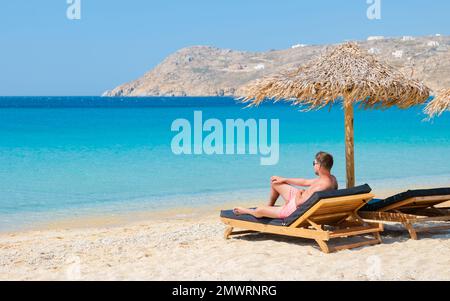 Junge Männer am Strand von Mykonos im Sommer mit Sonnenschirm und luxuriösen Liegestühlen, blaues Meer mit dem Berg am Elia Beach Mikonos Greece. Stockfoto