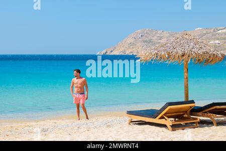Junge Männer am Strand von Mykonos im Sommer mit Sonnenschirm und luxuriösen Liegestühlen, blaues Meer mit dem Berg am Elia Beach Mikonos Greece. Stockfoto