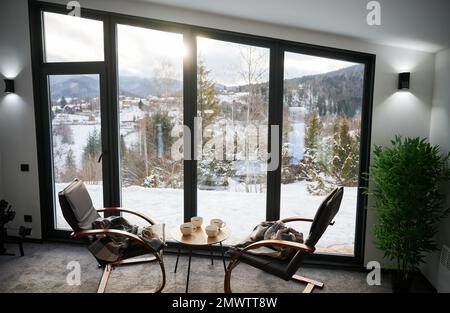 Gemütliches Zimmer mit kleinem Couchtisch und Sesseln vor einem großen Panoramafenster. Vorderansicht von zwei Holzstühlen und Tisch vor einem großen Fenster mit verschneiter Berglandschaft. Urlaubskonzept. Stockfoto