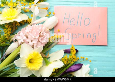 Rosafarbene Karte mit den Worten „HALLO FRÜHLING“ und frischen Blumen auf hellblauem Holztisch, flach liegend Stockfoto