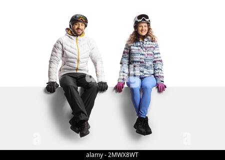 Junger Mann und junge Frau, die Winterjacken und Helme tragen und auf einer leeren Platte sitzen, isoliert auf weißem Hintergrund Stockfoto