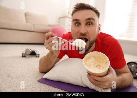 Fauler junger Mann, der Eis isst, anstatt zu Hause zu trainieren Stockfoto