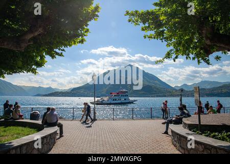 Touristen spazieren entlang des Seeufers von Varenna an einem sonnigen Tag mit blauem Himmel im Frühling. Panoramablick auf den See von Varenna, die Berge und A Stockfoto