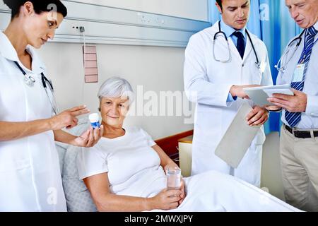 Ich überprüfe ihre Medikamente. Drei Ärzte, die einen älteren Patienten betreuen, der sich in einem Krankenhausbett befindet. Stockfoto