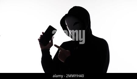 Anonymer Hacker und Gesichtsmaske mit Smartphone in der Hand. Mann in schwarzem Kapuzenhemd, der ein Mobiltelefon auf weißem Hintergrund hält und benutzt. Repräsentiere Cyber cr Stockfoto