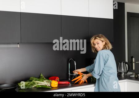 Fröhliche junge Frau wäscht Karotten in der Nähe von frischem Gemüse in der Küche, Stock Image Stockfoto