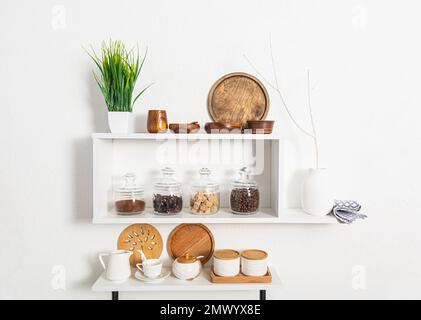 Ein Set aus Holz- und Keramikschalen, glasgefüllten Gläsern mit Großprodukten auf weißen Küchenregalen an strukturierten Wänden. Kompakter Stauraum. Elegantes Design Stockfoto