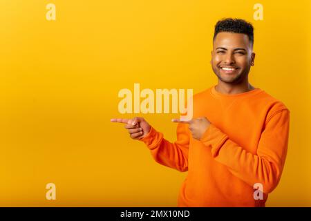 Porträt eines angenehm aussehenden indianers mit orangefarbenem Pullover, der mit den Fingern auf den Kopierbereich zeigt und einen Bereich für Werbung zeigt. Innenstudio-Aufnahme isoliert auf gelbem Hintergrund Stockfoto