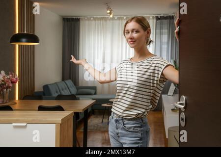 Fröhliche Frau, die die Haustür öffnet und Freunde einlädt. Junge Frau begrüßt Besucher in ihrem Haus. Stockfoto