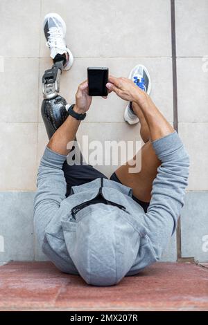 Draufsicht eines anonymen Sportlers mit Beinprothesen mit einem Mobiltelefon mit schwarzem Bildschirm während einer Pause in der Fitnessarbeit auf einem Gehweg nahe der Wand Stockfoto