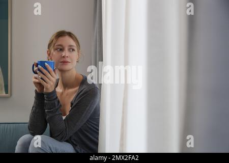 Entspannte junge Frau, die eine Tasse Kaffee im Wohnzimmer genießt, entspannender Selbstpflegetag zu Hause Konzept. Stockfoto