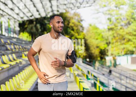 Junger hispanischer Mann mit Kopfhörern, der morgens im Stadion läuft, ein Mann, der Musik und Hörbücher anhört, Online-Podcasts über die App am Telefon, Läufer lächelt und mit dem täglichen Training zufrieden ist. Stockfoto