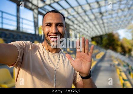 Sportläufer im Stadion, der Selfie-Fotos macht und mit Freunden per Videogespräch spricht, hispanischer Mann, der in die Kamera schaut und einen lächelnden Sport-Blog aufnimmt, junger Mann nach aktivem Training. Stockfoto
