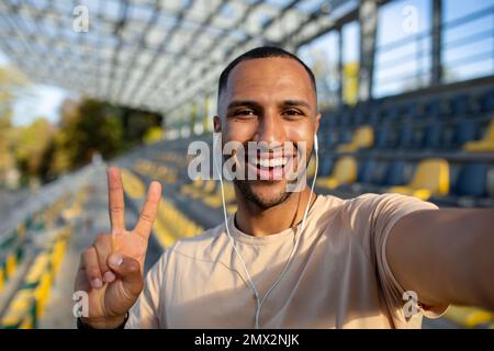 Sportläufer im Stadion, der Selfie-Fotos macht und mit Freunden per Videogespräch spricht, hispanischer Mann, der in die Kamera schaut und einen lächelnden Sport-Blog aufnimmt, junger Mann nach aktivem Training. Stockfoto