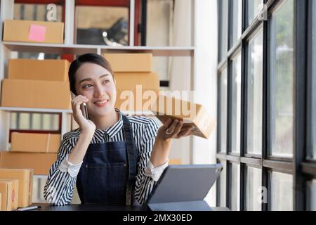 Eine asiatische Frau telefoniert mit einem Kunden, um eine Bestellung zu bestätigen. Sie besitzt einen Online-Shop, packt und versendet über einen privaten Transport Stockfoto