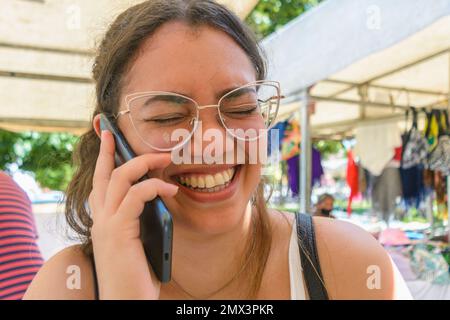 Die kleine venezolanische Latina-Frau, die Brille trägt und die Augen geschlossen hat, steht draußen und lacht am Telefon, während sie durch die Gegend geht Stockfoto