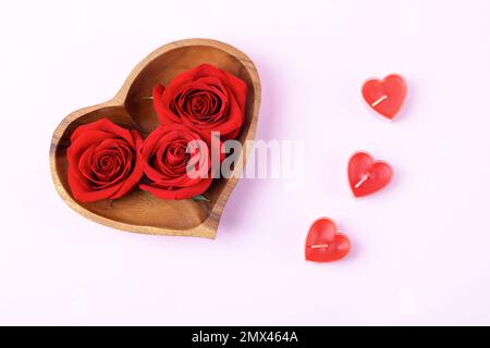 Drei rote Rosenknospen in einer Holzschüssel in Form eines Herzens mit drei roten Kerzen Herzen auf pinkfarbenem Hintergrund. Alles gute zum Valentinstag, zum Muttertag und Stockfoto