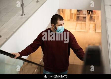 Mann mit Einwegmaske auf Rolltreppe im Einkaufszentrum. Gefährlicher Virus Stockfoto