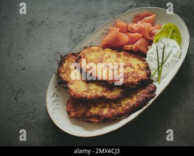 Blick von oben auf gebratene Kartoffelpfannkuchen und Lachs, serviert auf ovalem Teller neben Salatblättern und cremiger Sauce auf grauem Hintergrund Stockfoto