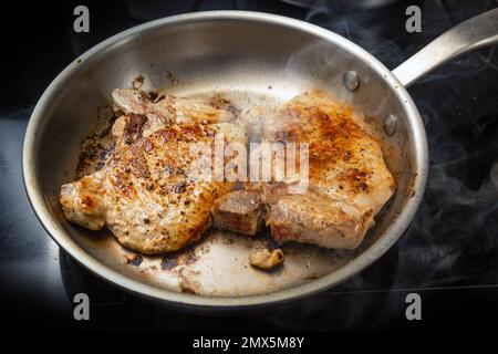 Zwei Schweinekoteletts werden in einer Bratpfanne aus Edelstahl auf einem schwarzen Kochfeld geröstet. Das Essen- und Kochkonzept, der ausgewählte Fokus, die geringe Schärfentiefe Stockfoto