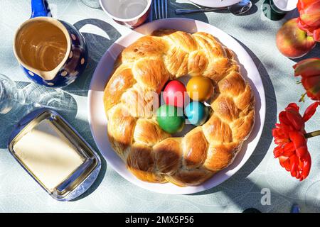 Traditionelles Ostergeflecht in Kranzform, gebacken aus einem leicht süßen Hefeteig mit bunten Eiern in der Mitte auf einem Tisch draußen in der Sonne, h Stockfoto