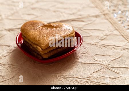 Ein herzförmiger Pfannkuchen in einer roten herzförmigen Untertasse auf einer Tischdecke aus Spitze - romantisches valentinstag-Konzept Stockfoto