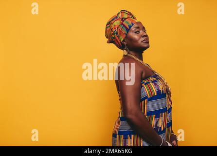 Stilvolle afrikanische Frau mit traditioneller Kleidung auf gelbem Hintergrund. Reife schwarze Frau in farbenfrohem Kente-Stoff und goldener Jewelle Stockfoto