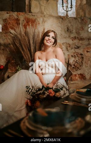 Wunderschöne Braut am Hochzeitstisch, reich dekoriert mit Blumen und Kerzen Stockfoto